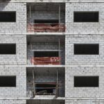 Fachada de un edificio en construcción, mostrando muros de bloques de concreto y balcones con malla de seguridad. Se observa un contenedor rojo y tuberías expuestas en la estructura.