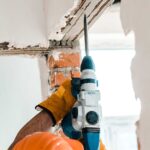 Trabajador utilizando un taladro eléctrico para realizar trabajos de demolición en una pared, con guantes de protección y casco anaranjado.